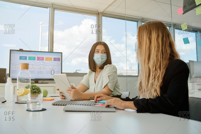 Serious young female coworkers in medical masks sitting at table with gadgets and papers and having conversation about business results during meeting in spacious office room