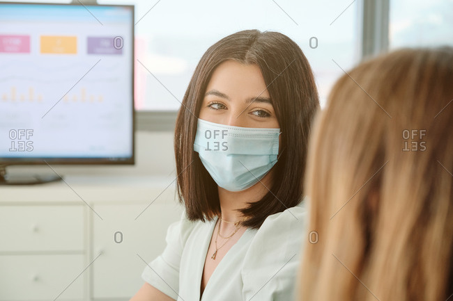 Serious young female coworkers in medical masks sitting at table with gadgets and papers and having conversation about business results during meeting in spacious office room