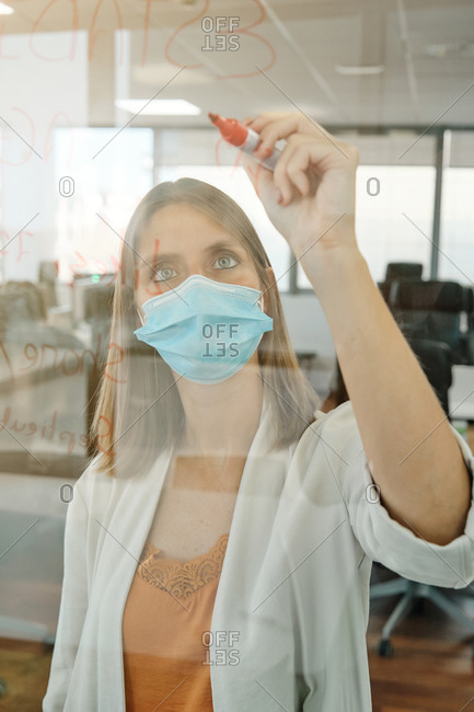 Young female member of business team in protective mask taking notes on glass wall while working with colleagues and searching for solution of business problem in contemporary office