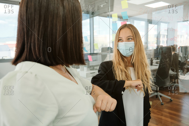Young female coworkers in medical masks bumping elbows while greeting each other in modern office during coronavirus pandemic