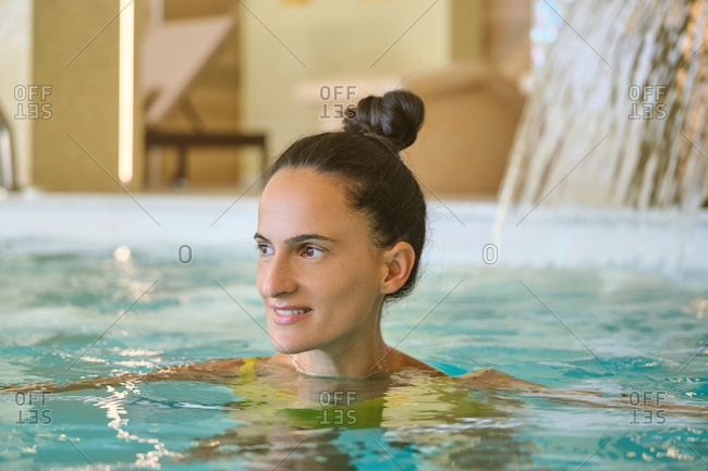 Positive young female getting hydrotherapy massage under waterfall during spa session in pool of luxury spa center in resort