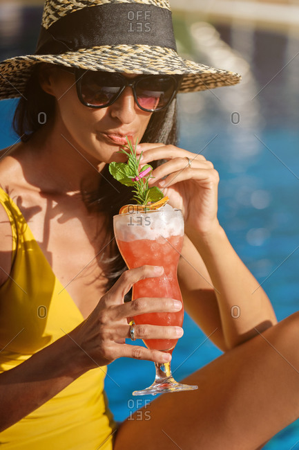 Young female in swimsuit and straw hat with sunglasses relaxing on lounge with glass of cold beverage while spending summer holidays in luxury resort