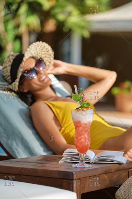 Side view of young female in swimsuit and straw hat with sunglasses relaxing on lounge with book and glass of cold beverage while spending summer holidays in luxury resort