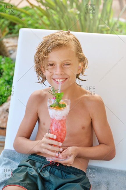 Cheerful preteen boy with wet curly hair sitting on chair and enjoying refreshing fruit drink while spending summer day at poolside in resort