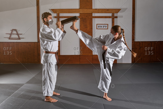 Woman with braid wearing protective mask and white kimono with a black belt attached kicking a man\'s mitten into the air