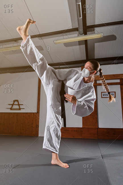 Woman with a white kimono and braid wearing a protective face mask with a black belt tied up kicking in the air in a martial arts gym