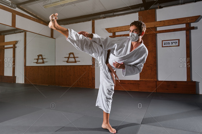 Woman with a white kimono and braid wearing a protective face mask with a black belt tied up kicking in the air in a martial arts gym
