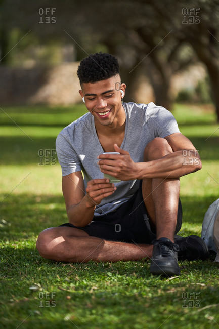 African American sportsman sitting on lawn with basketball and skate while relaxing after training and chatting on social media via mobile phone