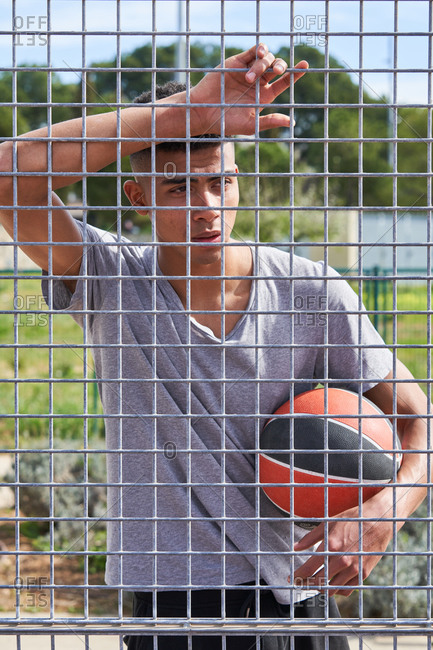 Calm African American player standing with basketball and leaning on metal grid on playground while looking away