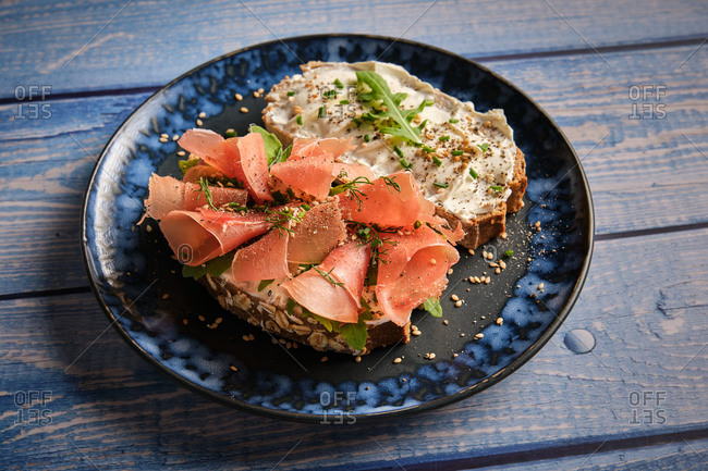 Appetizing toasts made with wholegrain bread with cream cheese and sliced ham garnished with fresh green herbs served on plate on wooden table for healthy breakfast
