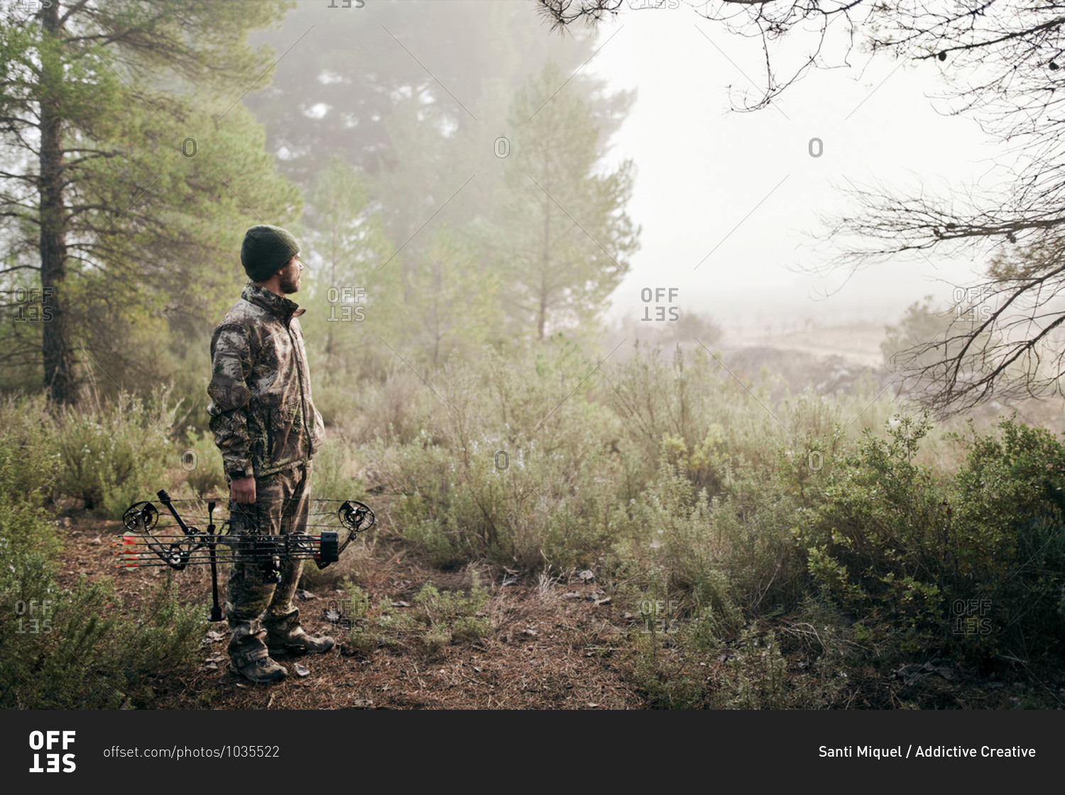Side view of man in camouflage standing with compound bow in forest and looking away during hunting