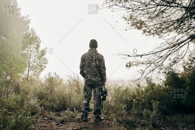 Back view of unrecognizable man in camouflage standing with compound bow in forest and looking away during hunting
