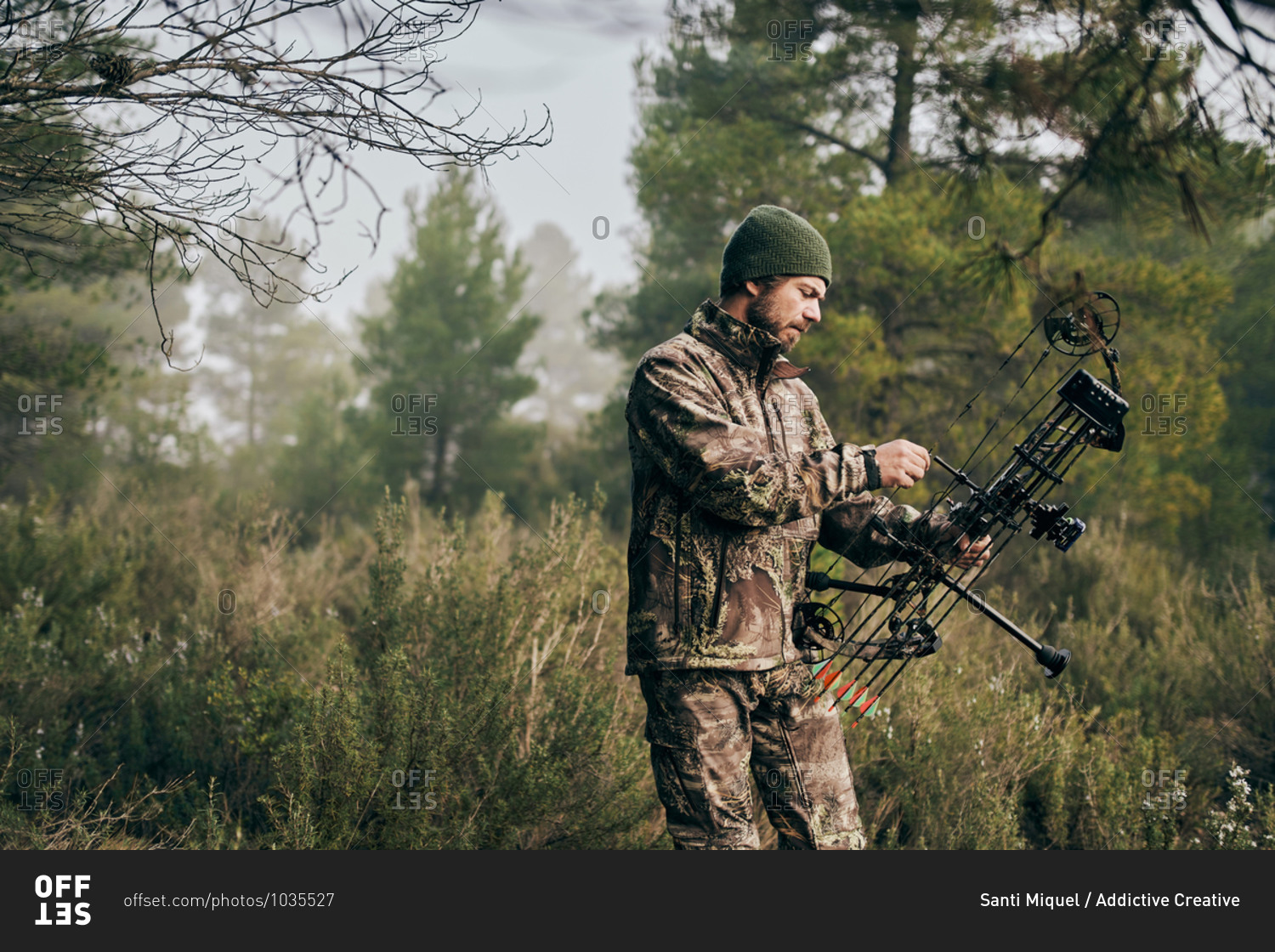Side view of serious male hunter adjusting compound bow with arrow while preparing for hunting in woods in autumn