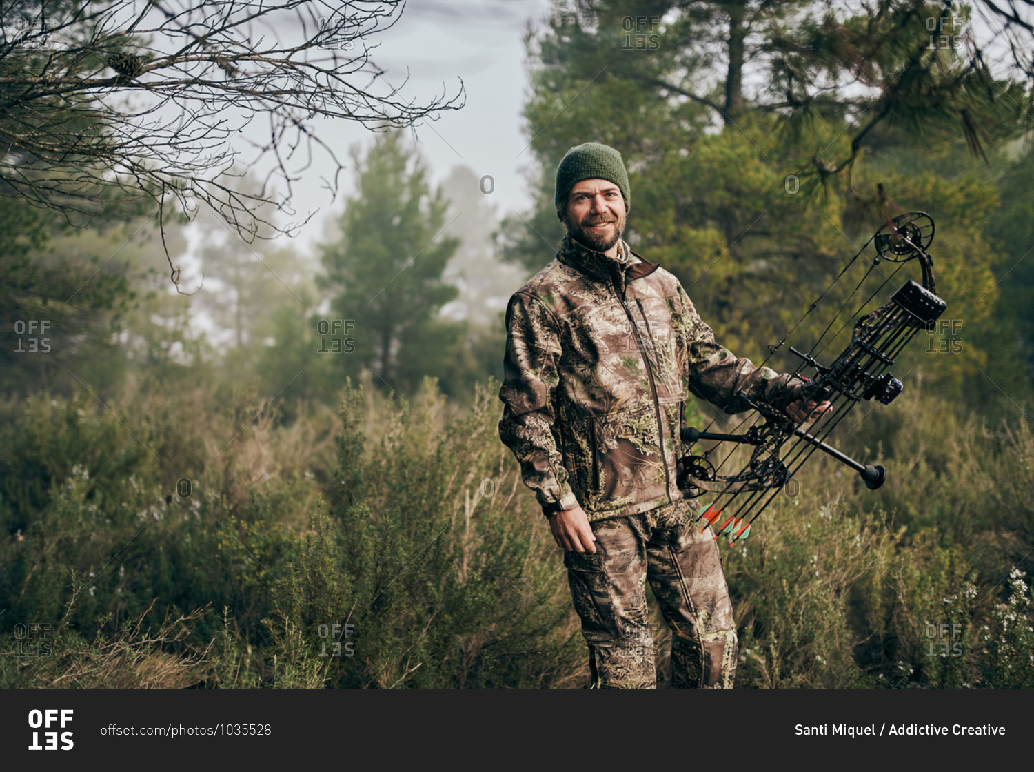 Cheerful man in camouflage standing with compound bow in forest and looking at camera during hunting
