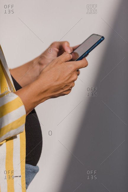 Side view of crop female expecting baby dressed in stylish striped jacket using smartphone while standing against white wall with sunlight and shadows