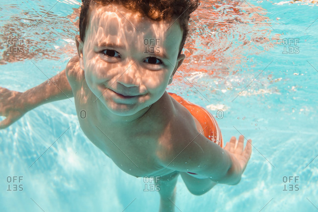 Underwater view of delighted kid swimming in clear water of pool and looking at camera