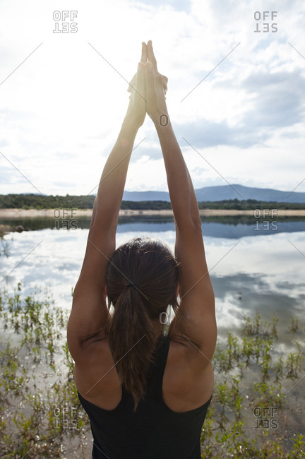 Back view of unrecognizable female in sportswear practicing Warrior asana with arms raised while doing yoga near lake