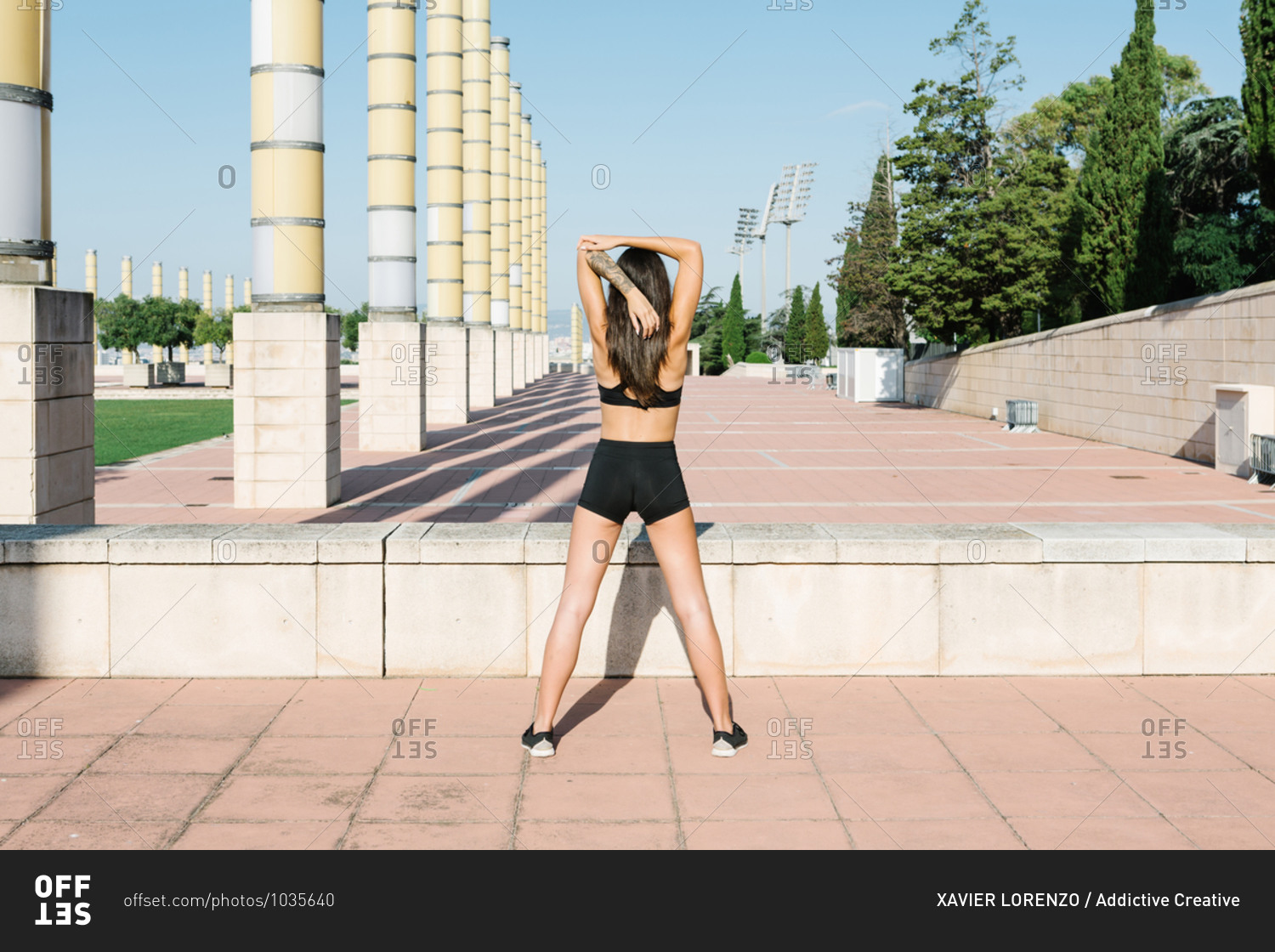 Full body back view of unrecognizable slim female in sportswear stretching shoulders while warming up before fitness workout on urban square