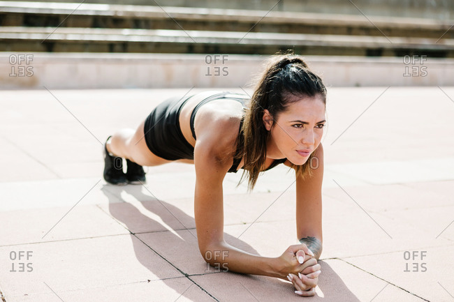 Full length of motivated strong fit female in sportswear doing forearm plank exercise during intense abs training on paved square in city