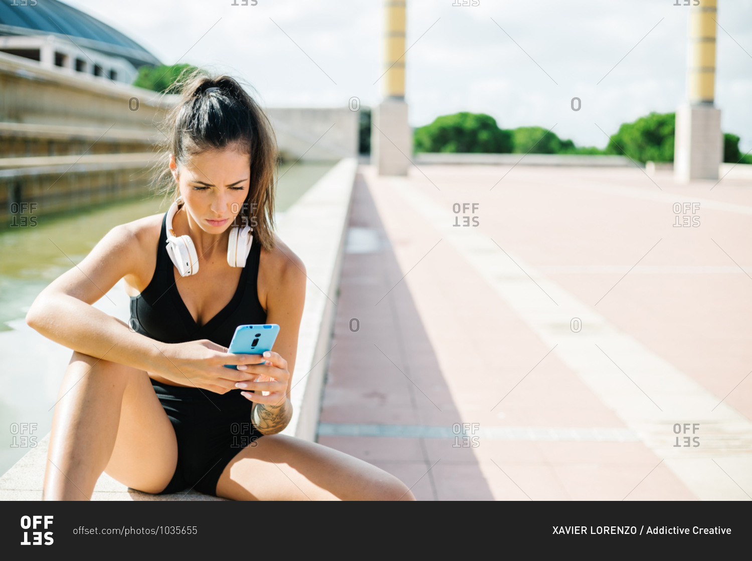 Fit female with tattoo on arm and headphones on neck dressed in black sports bra and shorts browsing mobile phone while sitting on stone border and relaxing after fitness workout on street