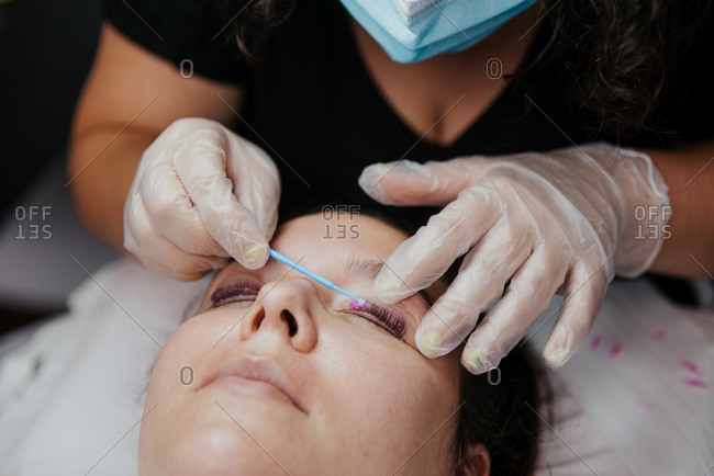 Closeup of crop unrecognizable cosmetician in latex gloves applying purple chemical solution on eyelid of female client during eyelashes lifting procedure in salon