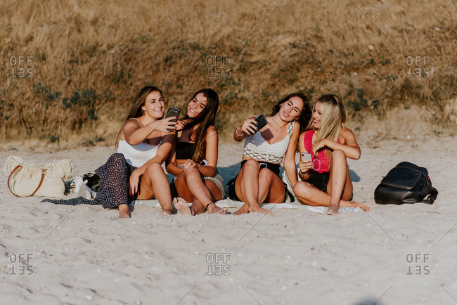 Group of smiling young ladies in casual clothes sitting on sandy beach and taking selfie with mobile phones while enjoying summer holidays together