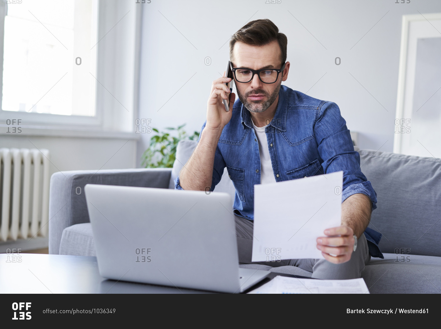 Serious man with documents and laptop on sofa talking on cell phone