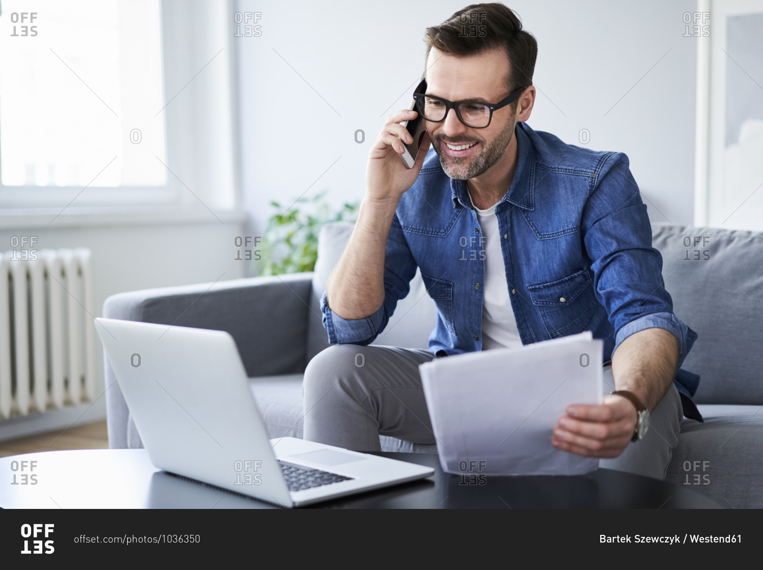 Smiling man with documents and laptop on sofa talking on cell phone