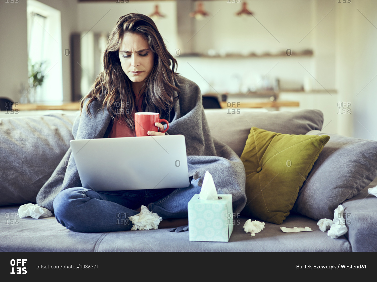 Sick woman sitting on sofa covered in blanket with cup of tea and laptop