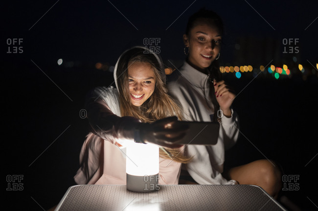Two female friends with led light on camping table at night taking a selfie