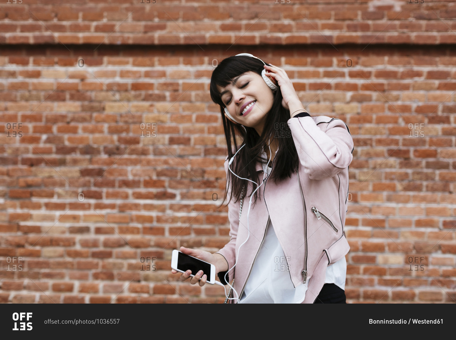 Smiling woman with cell phone listening to music on headphones at brick wall
