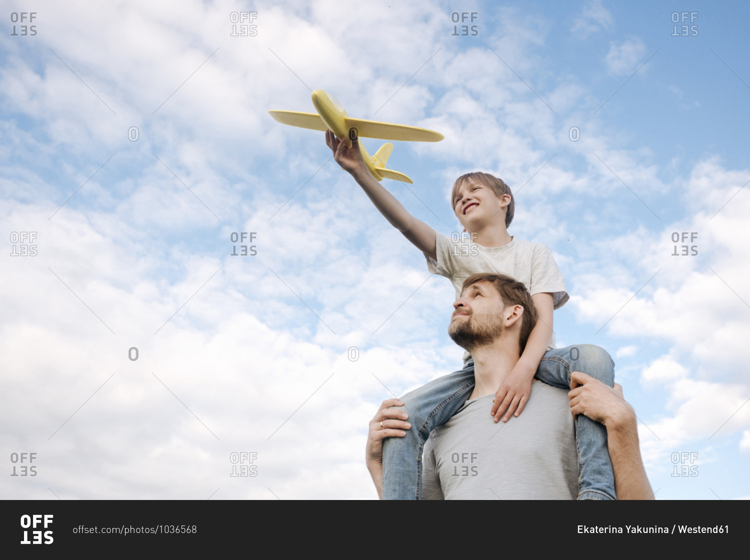 Man carrying son on shoulders playing with toy airplane against sky