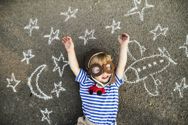 Portrait of smiling toddler wearing pilot hat and goggles lying on asphalt painted with airplane- moon and stars