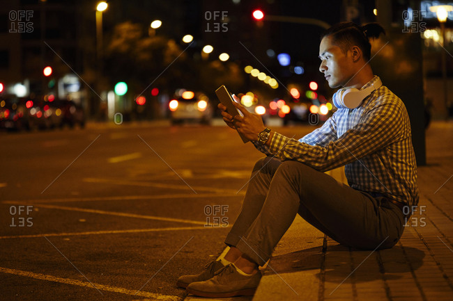 Young man sitting at roadside in the city at night using tablet