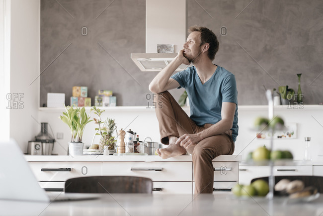 Smiling man sitting on kitchen counter looking at distance