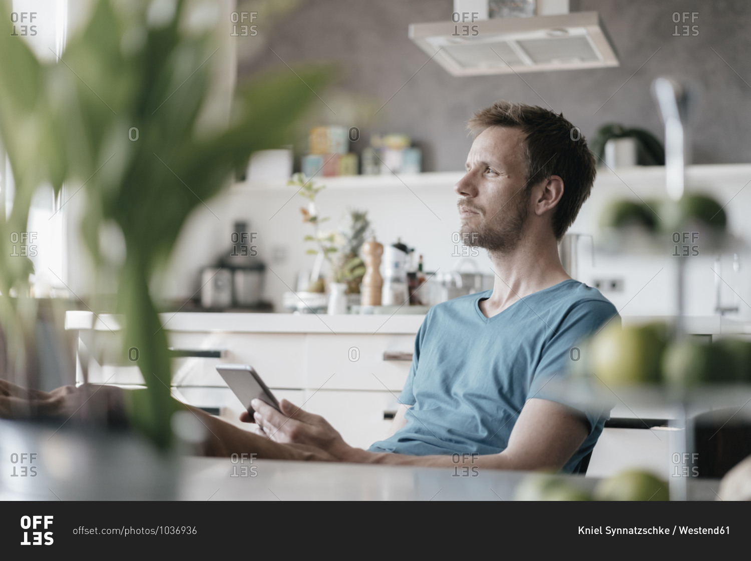 Serious man with tablet sitting in the kitchen with feet up looking at distance