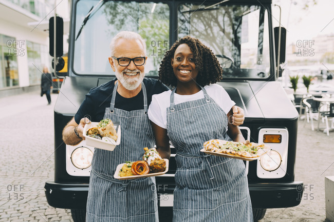 Smiling senior owner with assistant holding food plate while standing against commercial land vehicle