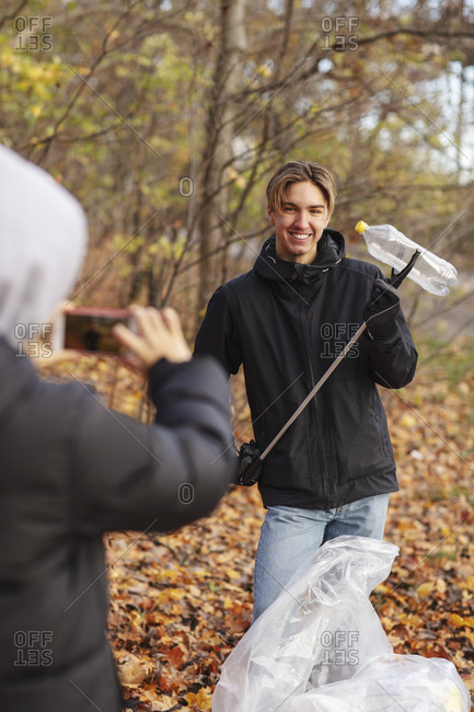 Teenager photographing friend with waste plastic bottle in park during autumn