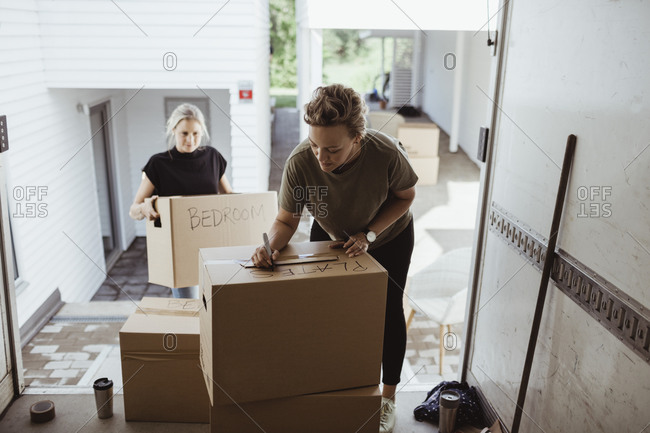 Woman holding box while female writing on cardboard box in moving van