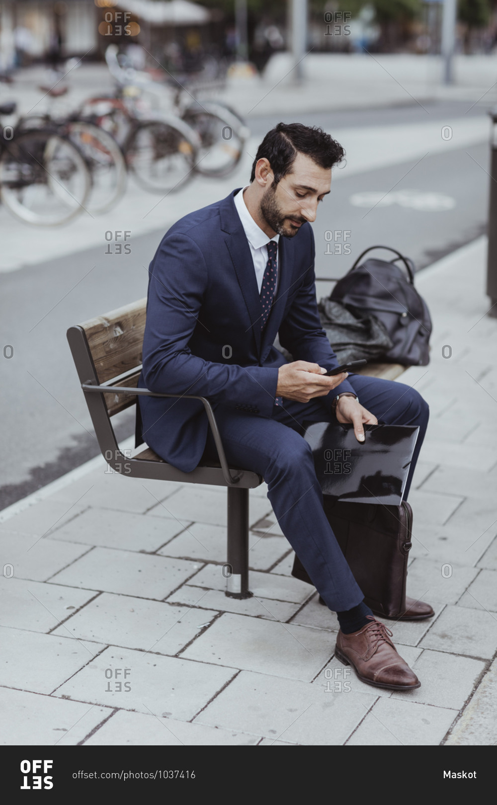 Entrepreneur with bag and file using smart phone while sitting on bench in city