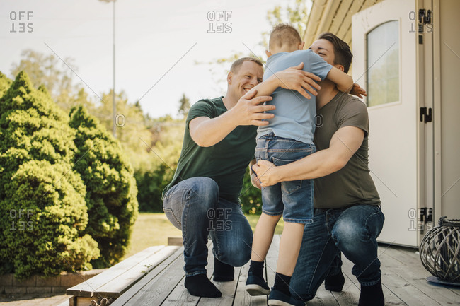 Homosexual fathers embracing son while crouching on patio during sunny day