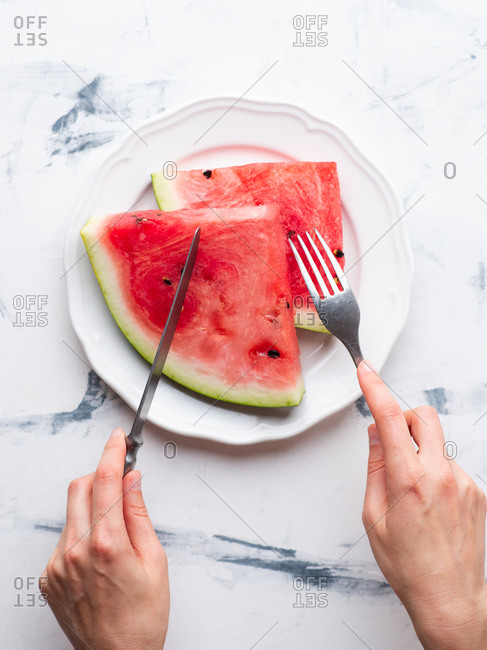 Sliced watermelon served on ceramic white plate over white background. Female hands holding fork and knife