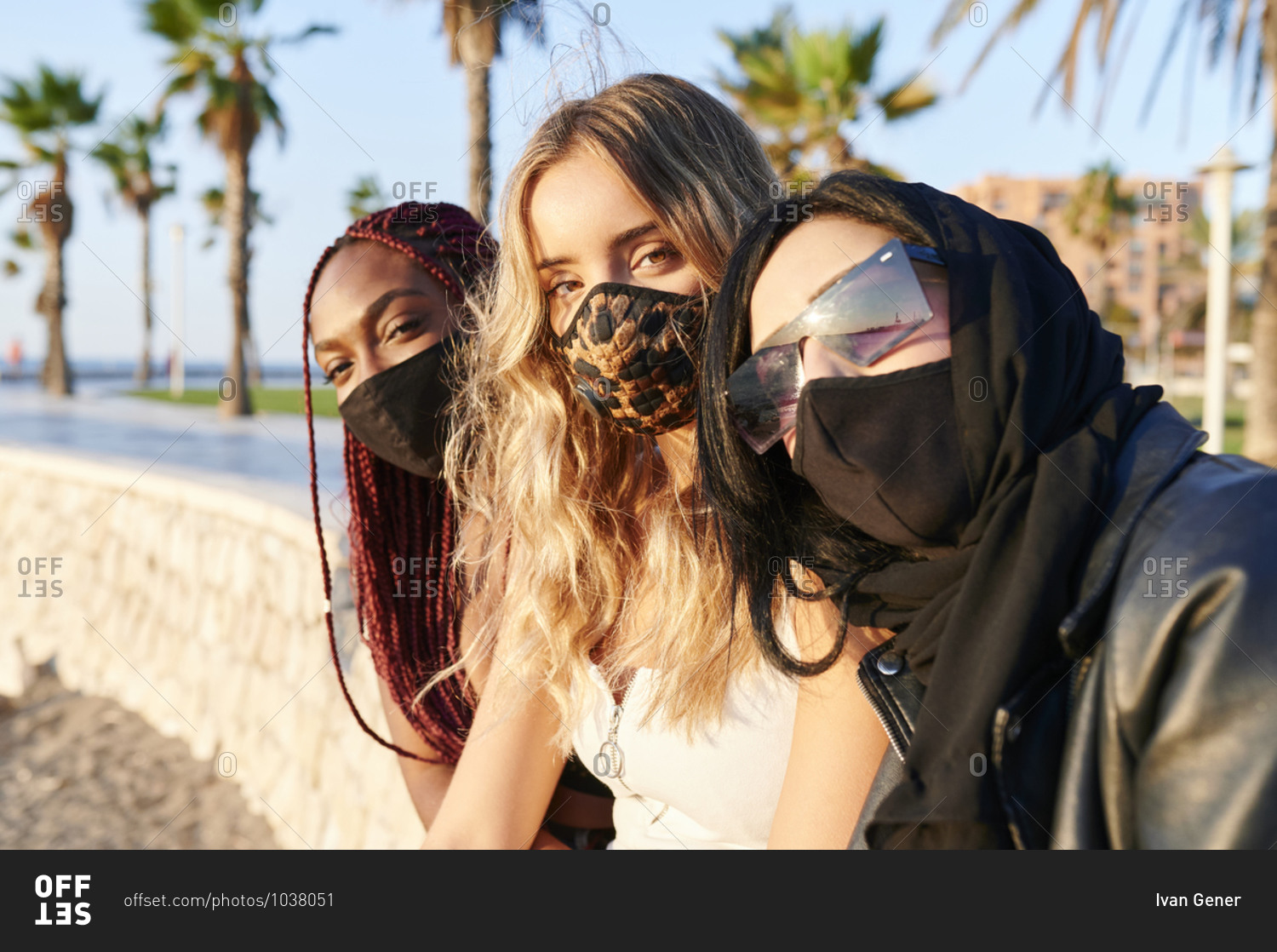 Diverse group of young female friends in face masks smiling from under face mask while sitting in a city park
