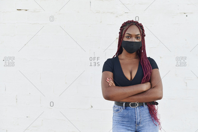 Young African woman standing with her arms crossed in front of a brick wall wearing a protective face mask