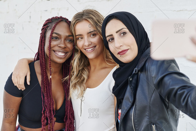 Three diverse young woman smiling and taking selfies together while standing outside
