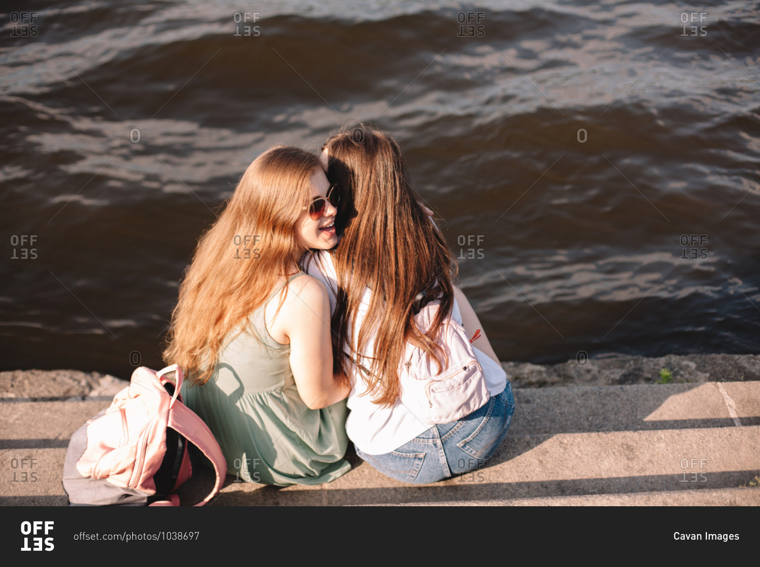 Young woman whispering into her girlfriend\'s ear sitting by river