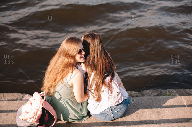 Young woman whispering into her girlfriend\'s ear sitting by river