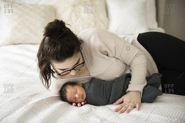 Hipster Millennial Mom Snuggles Swaddled Newborn on White Bed