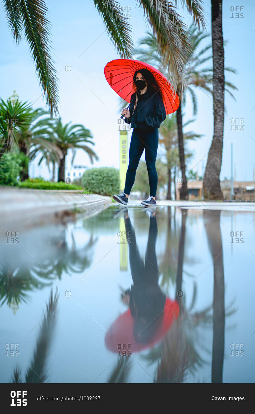 Woman with a mask and a red umbrella reflected on a puddle