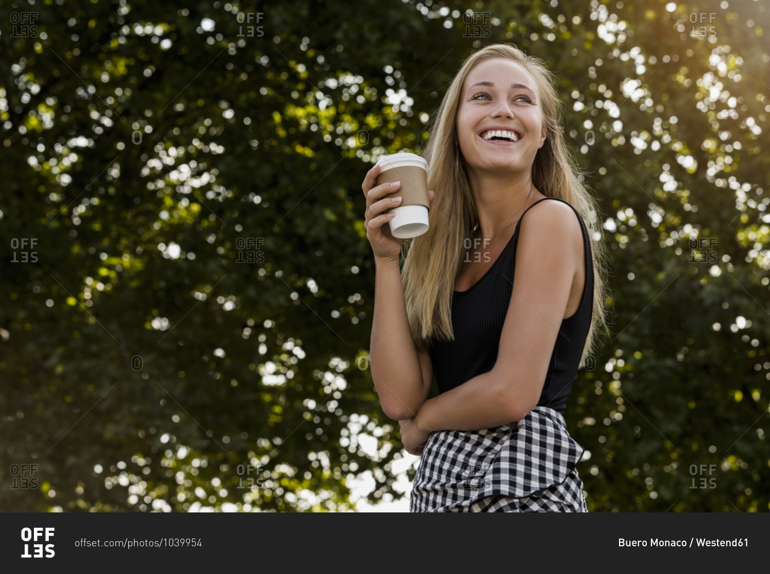 Cheerful young woman holding coffee cup while standing against trees in park